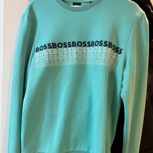Turkos sweatshirt från BOSS - Säljer en fräsch turkos sweatshirt från BOSS med tryckt logga i svart och vitt över bröstet. Tröjan har rund hals och långa ärmar, perfekt för chill dagar. Mjuk bomullskvalitet och relaxed passform. Snygg att matcha med jeans eller joggers.