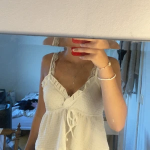 Vit omlott linnetopp med spets - Supersöt vit linnetopp med omlott och knyt framtill. Spetsdetaljer vid axelbanden och v-ringad hals ger en romantisk vibe. Lätt och luftig, perfekt för varma dagar. Volangkant upptill och ribbad struktur gör den extra charmig.