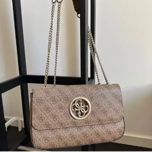 Beige axelväska från Guess med kedja - Jättefin Guess väska med dubbla axelband. Ca 28 x 19 x 6cm. Lite repig på märket i fram och en fläck i ena hörnet på insidan av väskan därav billigare pris. Köpt för ca 2000kr, Pris kan diskuteras!