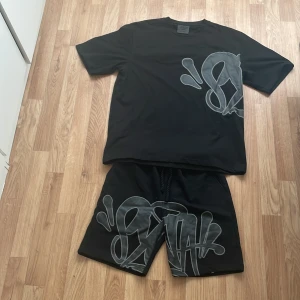 Svarta shorts och T-shirt från Syna world med graffiti -  (Både tröja och shorts). Svarta shorts från Syna world i storlek L med grått graffiti-tryck framtill. De har elastisk midja med snörning och en bakficka. Materialet känns som mjuk bomull eller polyester, perfekt för chill dagar. Snygg streetstil med unik design. (Inte blivit använda, problemet va att jag inte tyckte om de)