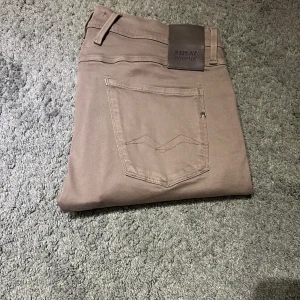 Replay Anbass Hyperflex beige jeans - Snygga Replay Anbass Hyperflex jeans i beige med slim fit och klassisk femficksdesign. Stretchigt material för extra komfort och rörelsefrihet. Diskret Replay-logga på fickan och bakre läderpatch. Perfekta för dig som gillar stilrena och bekväma jeans.