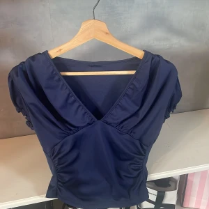 Mörkblå v-ringad blus med puffärm - Supersnygg mörkblå blus med v-ringning och puffiga korta ärmar. Snygga rynkade detaljer vid axlar och midja ger en cool siluett. Perfekt att styla med jeans eller kjol för en trendig look. Materialet känns mjukt och har lite stretch.