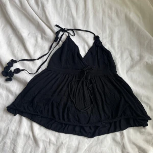 Svart linnetopp  - Supersöt svart linnetopp med v-ringad hals och knytband i nacken som har små tygrosor som detalj. Toppen har en flowy, volangliknande nederdel och markerad midja. Perfekt för dig som gillar en feminin och chill stil.