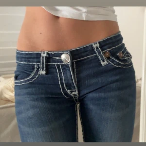 True Religion lågmidjade jeans  - Säljer ett par mörkblå jeans från True Religion med tydliga vita kontrastsömmar och klassiska U-formade bakfickor. Modellen är lågmidjad och har flare ben med snygga detaljer vid fickorna och en stor logopatch bak. Perfekt för dig som gillar statement-jeans! Dragkedjan är lite sönder men det går att fixa💕    Midjemått: 38cm