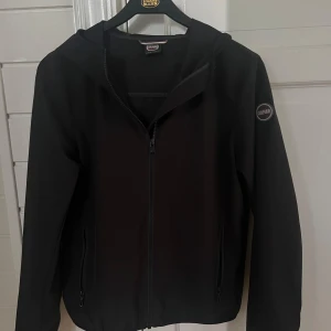 Svart vindjacka från Colmar med huva - Säljer en svart windbreaker från Colmar med stilren design och huva. Jackan har hel dragkedja framtill, två sidofickor med dragkedja och Colmar-logga på ärmen. Materialet är syntet och jackan har en normal passform med långa ärmar jackan har dessutom inga defekter. Storlek 16 years 172 cm
