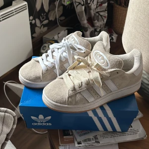 Beige adidas Campus sneakers strl 38 - Pris kan diskuteras!!! Säljer ett par fräscha adidas Campus sneakers i beige mocka med vita detaljer och klassiska tre ränder på sidan. Skorna har rund tå, platt sula och kommer med både vita och beige skosnören. Perfekt för dig som gillar clean och retro stil.