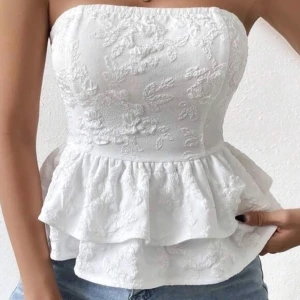 Vit blommig tubtopp med rosett - Superfin vit tubtopp med blommigt broderat mönster och volangdetaljer. Toppen har en stor rosett i ryggen och är axelbandslös med bandeauform. Perfekt för dig som vill ha en söt och trendig look till sommaren.