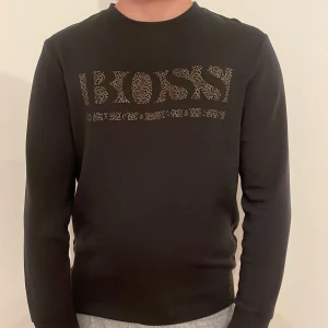 Svart BOSS tröja med guldtryck - Svart långärmad tröja från Hugo Boss i storlek S. Framsidan har ett stort BOSS-tryck i guldiga prickar som ger en lyxig vibe. Tröjan har rund hals och är gjord i mjukt bomullsmaterial, perfekt för dig som gillar stilrena och snygga plagg med lite extra attityd.