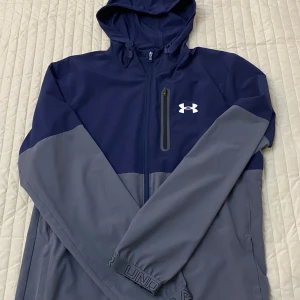 Blå och grå vindjacka Under Armour - Snygg tvåfärgad vindjacka från Under Armour i marinblått och grått. Jackan har huva, hel dragkedja framtill och en praktisk bröstficka med zip. Perfekt för dig som gillar sportig stil och vill ha något lätt och funktionellt.
