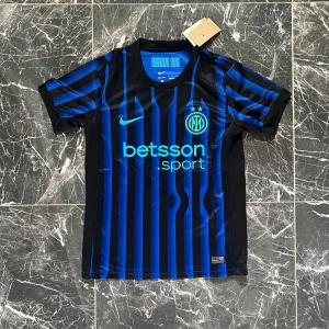 Inter Milan randig fotbollströja Nike - Snygg Inter Milan fotbollströja från Nike med klassiska blå och svarta ränder. Kortärmad modell med rund hals och tryckta loggor på bröstet. Tillverkad i lätt och ventilerande polyester, perfekt för match eller träning. Betsson Sport sponsortryck på framsidan.