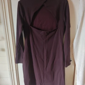 Mörklila klänning med öppen rygg - En mörklila klänning med lång ärm och en öppen, triangelformade ryggdetalj. Den är inte särskilt kort men slutar ovanför knäet. Den är ganska stretchig och tror att det är meningen att den ska sitta tätt intill kroppen. Men det gör man som man vill.