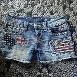 Miss Me jeansshorts med nitar och patch - Snygga blå jeansshorts från Miss Me med rå kant, slitningar och patchade detaljer i rött, vitt och blått. Shortsens framsida har nitar och broderade flaggmotiv, bakfickorna är dekorerade med nitar och coola Miss Me-loggor. Perfekt för dig som gillar edgy och unik stil.
