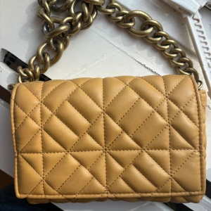 Quiltad beige axelväska från Zara - Säljer en quiltad beige/senapsgul axelväska från Zara med grov guldfärgad kedja som axelrem. Väskan har ett magnetlås och insidan är fodrad i svart tyg. Perfekt storlek för mobil, plånbok och småsaker. Stilren design med rutmönster på utsidan.