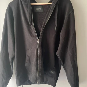 Svart hoodie från Replay med dragkedja - Svart hoodie från Replay med hel dragkedja framtill, huva med snörning och två fickor. Diskret Replay-logga på bröstet och mjukt bomullsmaterial. Perfekt för chill dagar och enkel att matcha med jeans eller joggers.