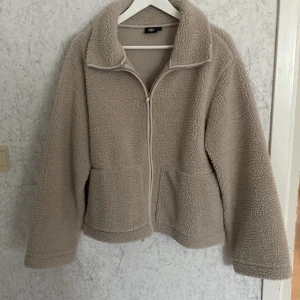 Beige teddyjacka från 157 - Mysig beige teddyjacka från 157 med fluffigt material och dragkedja framtill. Jackan har två stora fickor, hög krage och långärmade ärmar. Perfekt för att hålla sig varm och se chill ut under höst och vinter. Enkel, loose passform som ger en avslappnad vibe.