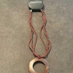 Brunt halsband från Freedom - Unikt halsband med flera rader små bruna pärlor och ett stort, runt hänge i mörkbrunt naturmaterial. Hänget har en cool, organisk känsla och pärlorna ger en bohemisk vibe. Perfekt för dig som vill sticka ut med accessoarer som känns personliga.  Står ej för frakt 
