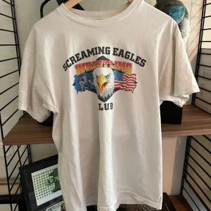 Amerikansk vintage-T-shirt - Vit Amerikansk vintage-T-shirt med tryck av en örn, amerikansk flagga och texten 'Screaming Eagles Wrestling Club' på bröstet. Klassisk rund hals och korta ärmar. Materialet är en mix av bomull och polyester, vilket gör den mjuk och skön att ha på sig.