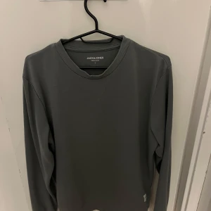 Mörkgrå sweatshirt från Jack & Jones - En stilren grön/grå sweatshirt från Jack & Jones Essentials. Tröjan har rund hals, långa ärmar och en enkel design utan mönster eller detaljer. Perfekt för dig som gillar en clean och avslappnad look.