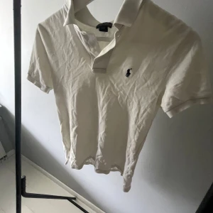 Vit piké från Polo Ralph Lauren - Snygg vit pikétröja från Polo Ralph Lauren med klassisk krage och korta ärmar. Tröjan har den ikoniska broderade loggan på bröstet och är gjord i mjuk bomull. Perfekt för dig som gillar stilrena och tidlösa plagg. 140/152
