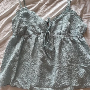 Ljusblå linnetopp från Nelly - Supersöt ljusblå linnetopp från Nelly med tunna axelband och volangdetaljer vid halsringningen. Toppen har en snygg knytning framtill och är gjord i ett krinklat, lätt material som känns luftigt. Banden är justerbara.