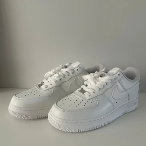 Nike Air Force 1 vita sneakers - Fräscha Nike Air Force 1 sneakers i helvitt skinn med klassisk låg siluett. Ikonisk design med perforerad tå, rundad sula och tydliga swoosh-loggor på sidorna. Skorna har snörning och platt sula, perfekt för dig som gillar cleana och stilrena sneakers.