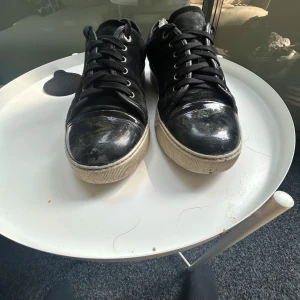 Svarta sneakers med mockadetaljer - Säljer ett par svarta sneakers med snygga mockadetaljer på sidorna och blankare material på tån. Skorna har rund tå, snörning och en vit platt sula som ger en cool kontrast. Perfekt för dig som gillar stilrena och klassiska sneakers med lite edge.