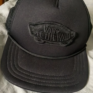 Svart Vans truckerkeps med mesh - Säljer en svart Vans truckerkeps med broderad 'Off The Wall'-logga framtill. Kepsen har böjd skärm, mesh på baksidan och justerbar snapback. Stilren och enkel design som passar till streetwear och skate.