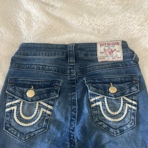 True Religion Becca Bootcut Jeans vita fickor  - Säljer ett par snygga blå jeans från True Religion, modell Becca med låg midja och bootcut. Jeansen har ikoniska broderade fickor med vita detaljer och guldiga knappar. Materialet är klassisk jeans med  väldig stretch för skön passform. Perfekt för dig som gillar statement-plagg! Dom är använda 2 gånger. Som nya jeans! Väldigt snygga. Dom går inte att köpa längre!