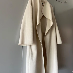 Beige kappa från Bianca x Nelly - Säljer en stilren beige/vit kappa från Bianca x Nelly i storlek 34. Jackan har en snygg omlottkrage och är tillverkad i mjukt material med lite struktur. Perfekt för dig som gillar minimalistisk och clean look. Passar dig som vill ha en klassisk vibe.                                         Har inga defekter men skulle behöva en tvätt🤍
