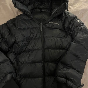 Svart Peak Performance dunjacka - Säljer en svart dunjacka från Peak Performance med huva och snygga detaljer. Jackan har broderad logga på bröstet och är quiltad med dragkedja framtill. Perfekt för kalla dagar och har en cool, puffig look som sticker ut.