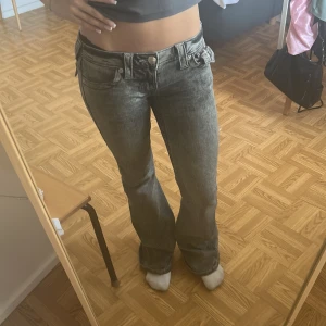 Grå bootcut jeans från True Religion - Säljer ett par gråa bootcut jeans från True Religion med låg midja och snygg tvättad look. Jeansen har detaljerade bakfickor med silverknappar och breda bälteshällor. Finns inga defekter alls men säljer pga att de inte används längre!