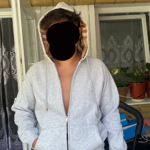 Grå Burberry hoodie med rutigt foder - Säljer en grå hoodie från Burberry med klassiskt rutigt foder i huvan. Tröjan har dragkedja, snörning vid huvan och två fickor framtill. Tillverkad i 100% bomull och har snygga metall-detaljer på snöret och dragkedjan. Perfekt för dig som gillar stilren design med lyxig touch.