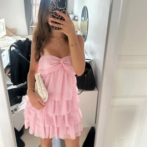 Rosa volangklänning - Supersöt rosa kortklänning med flera lager volanger och en snygg rosettdetalj framtill. Endast använd en gång så är i jättefint skick💕💕