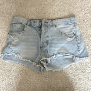 Ljusa jeansshorts från Pull&Bear - Säljer ett par ljusblå jeansshorts från Pull&Bear i storlek 36. De har hög midja, slitna detaljer och fransig kant nedtill för en chill vibe. Klassisk femficksmodell med knapp och dragkedja framtill. Perfekta för varma dagar!