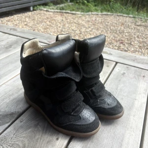 Svarta sneakers, Isabel Marant  - Säljer ett par svarta sneakers från Isabel Marant med inbyggd kilklack. Skorna har en cool design med breda kardborreband, rund tå och platt gummisula i beige. Materialet är en mix av mocka och skinn, vilket ger en lyxig känsla.❤️