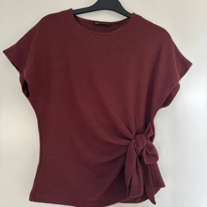 Mörkröd topp med knyt i sidan - Supersnygg mörkröd topp från Zara med rund hals och korta ärmar. Den har en cool knyt-detalj i sidan som ger extra stilpoäng. Materialet är mjukt och stickat, perfekt för att lyfta din outfit med en enkel twist.