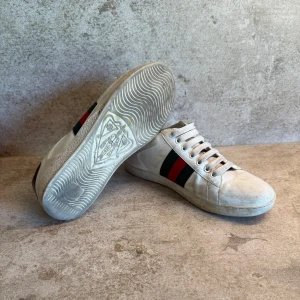 Gucci Ace sneakers med broderad bi - Gucci Ace sneakers med klassiska gröna och röda ränder på sidan samt broderad guldfärgad bi. Baksidan har metallicröd och metallicgrön häl med Gucci-logga. Platt sula med Gucci-mönster och snörning framtill. Perfekt för dig som vill sticka ut med lyxig streetstyle.