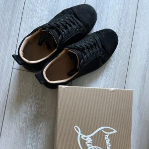 Svarta sneakers från Christian Louboutin - Snygga svarta sneakers från Christian Louboutin med klassisk röd sula och svart mocka på ovandelen. Skorna har rund tå, snörning och en stilren design som passar till många olika outfits. Insidan är ljusbeige och de levereras med originalkartong.