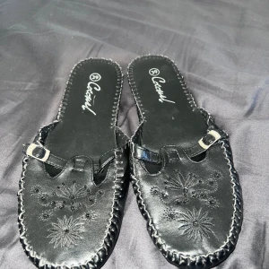 Svarta sandaler med broderi från Casual - Svarta sandaler från Casual med dekorativt broderi och små spänndetaljer framtill. Skorna har en platt sula och öppet hälparti