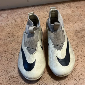 Nike Mercurial Air Zoom fotbollsskor - Nike Mercurial Air Zoom fotbollsskor i vit och grå med svarta detaljer och stor Swoosh-logga. Skorna har en strömlinjeformad design, täckt snörning och dragflik bak för enkel påtagning. Ovandelen är tillverkad i syntetmaterial för bra passform och grepp på planen.