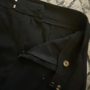 Svarta kostymbyxor med hög midja - Snygga svarta kostymbyxor med hög midja och raka ben. Byxorna har klassisk stängning med dragkedja, knapp och hyska, vilket ger en clean look. Perfekta för dig som gillar stilrena och tidlösa plagg. Materialet känns mjukt och har en lätt stretch.