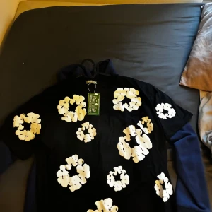 Svart t-shirt med vita blommor - Svart t-shirt från Wacko Maria med stora vita blommor tryckta över hela framsidan. T-shirten har rund hals och korta ärmar, perfekt för dig som gillar coola och unika prints. Materialet är mjuk bomull som känns skönt mot huden. Är även stor på en 