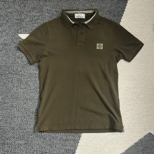 Olivgrön Stone Island piké t-shirt - Snygg olivgrön piké från Stone Island med klassisk krage och korta ärmar. T-shirten har vit och svart randig detalj på kragen och ett broderat Stone Island-märke på bröstet. Tillverkad i mjuk bomull som känns skön mot huden.