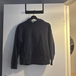 Mörkblå hoodie från Polo Ralph Lauren - Snygg mörkblå hoodie från Polo Ralph Lauren med klassisk röd logobrodyr på bröstet. Tröjan har huva, känguruficka och långa ärmar. Perfekt för chill dagar och enkel att matcha med jeans eller joggers.