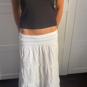 Vit långkjol med smock i midjan - Superfin vit långkjol med bred smock i midjan och volangiga lager. Kjolen är luftig och har en avslappnad passform, perfekt för varma dagar. Tillverkad i ett lätt tyg som känns skönt mot huden.