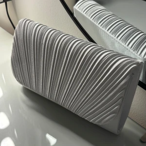 Silver vit  kuvertväska med veckad design - Snygg silver/vit färgad kuvertväska med stilrena veckade detaljer som ger en lyxig känsla. Perfekt storlek för det viktigaste och har en elegant form som passar till alla tillfällen. Materialet har en glansig finish som verkligen sticker ut. Endast använd vid ett tillfälle. 