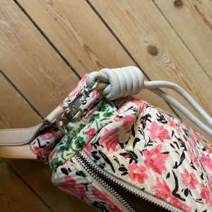 Blommig axelväska från Desigual - Axelväska från Desigual med blommigt mönster i rosa, grönt, gult och svart på beige bakgrund. Väskan har quiltad yta, flera dragkedjor och en justerbar ljus axelrem. Materialet är bomull och detaljerna är i silverfärgad metall. Perfekt för dig som gillar färg och unika accessoarer.