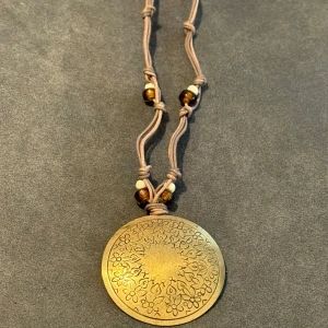 Boho halsband med stor guldbrun medaljong - Unikt halsband med stor rund medaljong i guldton med ingraverat blommönster. Hängs i en brun snodd med knutar och pärlor i brunt och beige. Perfekt för dig som gillar boho-stil och vill sticka ut med ett statement-smycke. Står ej för frakt 