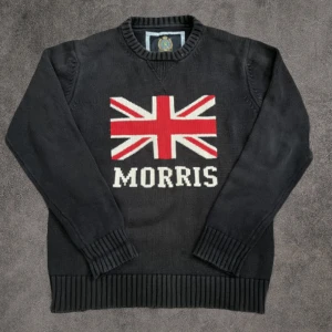 Svart stickad tröja Morris L - Svart stickad tröja från Morris i storlek Large med Union Jack-flagga och Morris-logga på bröstet. Tröjan har rund hals, ribbade muddar och nederkant. Materialet är mjukt och skönt, perfekt för kyliga dagar. Snygg och klassisk design med brittisk touch.