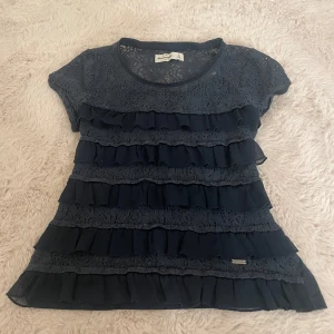 Mörkblå blus med spets från Abercrombie - Supersöt mörkblå blus från Abercrombie med korta ärmar, spets upptill och flera lager volanger framtill. Blusen har rund halsringning och detaljer i spets både fram och bak. Perfekt för dig som gillar romantisk och feminin stil. Hör gärna av er vid frågor osv 💗